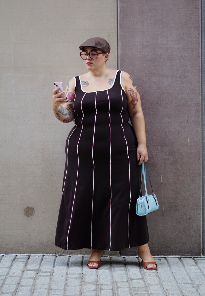 Robe maxi noire avec des rayures verticales roses, bretelles épaisses et encolure ronde. Portée avec des sandales à talons brunes et tenant un sac à main bleu clair.