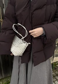 Άτομο με σκούρο καφέ φουσκωτό μπουφάν και γκρι πλισέ φούστα κρατάει μικρή λευκή υφαντή τσάντα με λαβές σε σχήμα θηλιών.