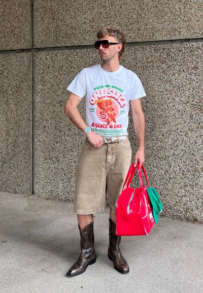 Camiseta gráfica blanca con estampado de pizza, pantalones cortos caqui, botas de vaquero marrones, bolsas rojas y verdes, de pie contra una pared texturizada.