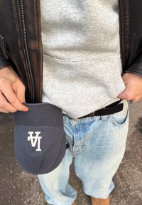 Sötétkék sapka fehér logóval, szürke kapucnis pulóver ellen tartva. A személy világoskék farmert és fekete bőrdzsekit visel.