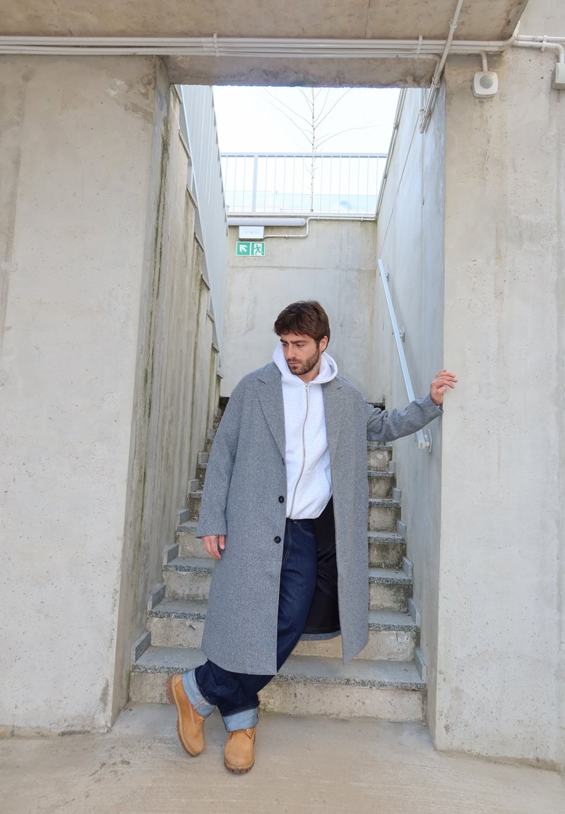 Man in a gray overcoat, white hoodie, dark jeans, and tan boots leaning on a handrail at a concrete stairwell with an exit sign above.