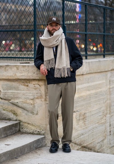 Hombre de pie contra una pared de piedra, vistiendo una chaqueta oscura, una bufanda beige con flecos, pantalones claros y zapatos negros, con una gorra de béisbol.