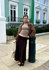 Brązowa zamszowa kurtka, beżowy golf i ciemnobrązowa spódnica maxi. Czarne buty i beżowa torebka. Zielony słupek z białym napisem w tle.