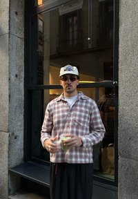 Homme portant une chemise en flanelle à carreaux rose et blanc, tenant une boisson verte, avec une casquette plate et des lunettes de soleil foncées. Debout près d'une fenêtre.