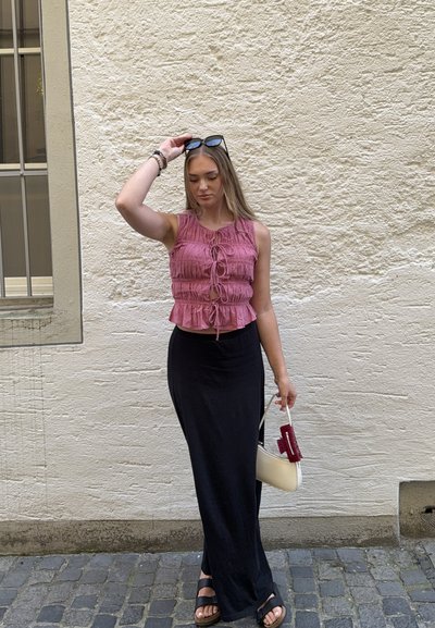 Top corto rosa con volantes y lazos en la parte delantera, falda maxi negra ajustada y sandalias negras. Bolso blanco con acentos rojos. El fondo presenta una pared texturizada.