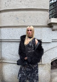 Personne blonde portant une veste noire en fourrure par-dessus une robe camouflage, tenant un sac noir, debout contre un mur en pierre texturé en plein air.