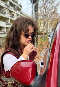 Sarkana transportlīdzekļa sānu spogulī atspoguļojas sieviete ar bordo vestīti un baltu topu, kas pilsētas vidē uzklāj kosmētiku no rozā tūbiņas.