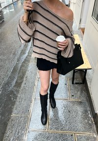 Helles beiges Strickoberteil mit schwarzen Streifen, schulterfreies Design, kombiniert mit einem schwarzen Rock, hohen schwarzen Lederstiefeln und einer Kaffeetasse in der Hand.
