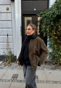 Brown suede jacket with a button front, paired with a black scarf and jumper. Grey wide-leg jeans complete the outfit. Green foliage in the background.