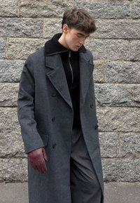 Grijze dubbelgeknoopte overjas met een geribbelde kraag, gedragen over een zwart vest met rits, gecombineerd met donkere broek en bordeauxrode leren handschoenen.