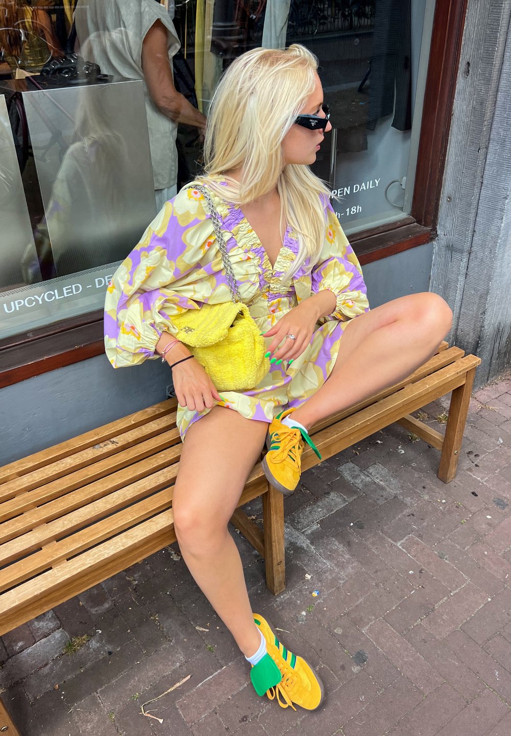 Φλοράλ φόρεμα σε κίτρινο και λιλά χρώμα με ριχτό λαιμό; φωτεινές κίτρινες υφασμάτινες τσάντες; κίτρινα αθλητικά παπούτσια με πράσινες και μπλε λεπτομέρειες.