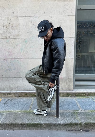 Hombre con chaqueta negra, gorra y gafas estira la pierna en la acera junto a un poste de metal frente a un fondo de pared de piedra.