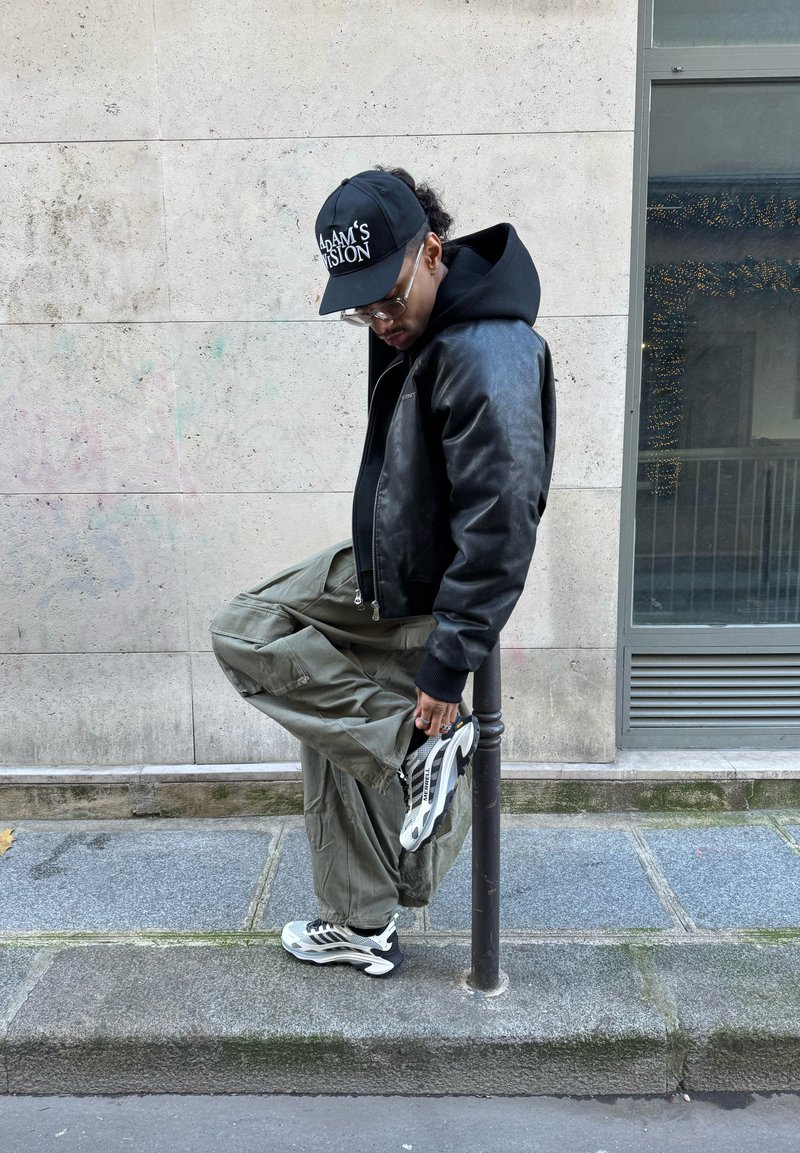 Mann iført svart jakke, caps og briller strekker ut benet på fortauet ved siden av en metallstolpe mot en bakgrunn av steinmur.