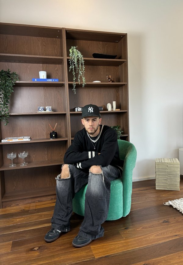 Green upholstered armchair, young man in black sweater and baggy jeans seated. Wooden shelves with decorative items and plants in background.