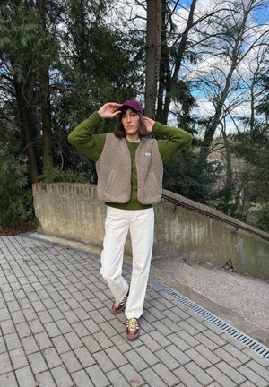 Persona ajustándose una gorra morada, con suéter verde, chaleco de forro polar beige, pantalones blancos y zapatillas moradas, de pie en un camino pavimentado cerca de árboles y escaleras.