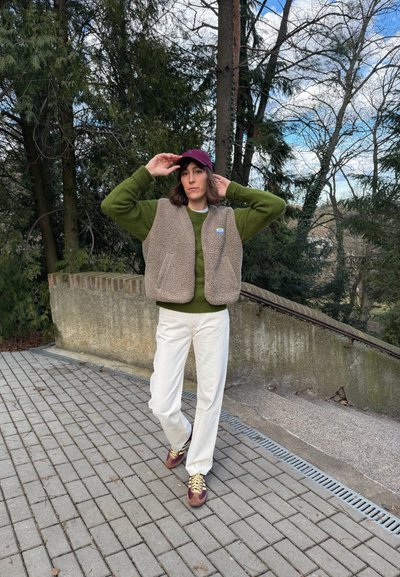 Persona ajustándose una gorra morada, con suéter verde, chaleco de forro polar beige, pantalones blancos y zapatillas moradas, de pie en un camino pavimentado cerca de árboles y escaleras.
