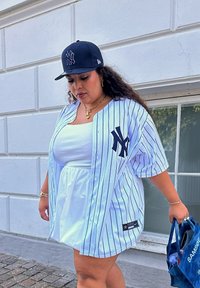 Λευκή και μπλε ριγέ μπλούζα μπέιζμπολ με λογότυπο, φορεμένη πάνω από ένα λευκό φόρεμα. Μαύρο καπέλο και κρατώντας μια μπλε τσάντα αγορών.