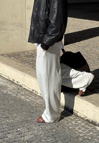 Giacca bomber nera in pelle, pantaloni della tuta grigi, scarpe slip-on marroni. In piedi su un pavimento di pietra con una borsa nera accanto.