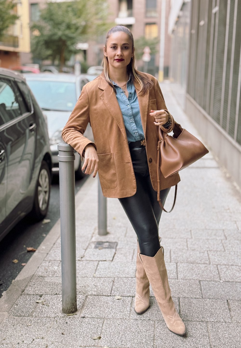 Καφέ σουέτ σακάκι πάνω από τζιν πουκάμισο, μαύρο δερμάτινο κολάν και μπεζ χαλαρό μποτάκι. Καφέ τσάντα και αστική σκηνή.