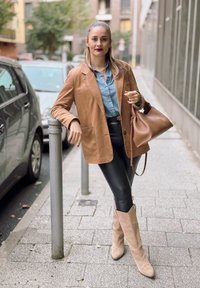 Καφέ σουέτ σακάκι πάνω από τζιν πουκάμισο, μαύρο δερμάτινο κολάν και μπεζ χαλαρό μποτάκι. Καφέ τσάντα και αστική σκηνή.