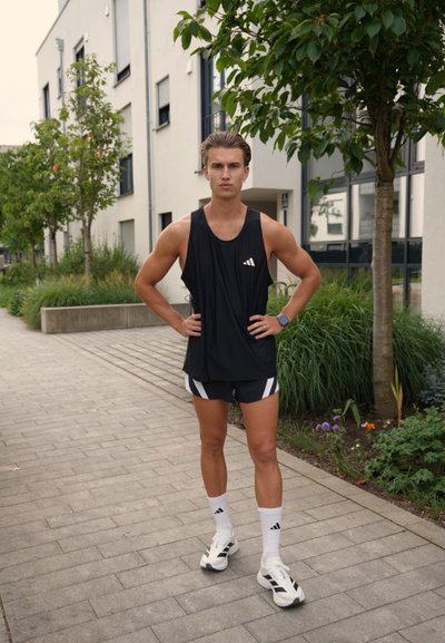 Camiseta de tirantes negra deportiva con logo blanco, pantalones cortos negros con rayas blancas, calcetines blancos y zapatillas con un diseño. Entorno exterior con vegetación.