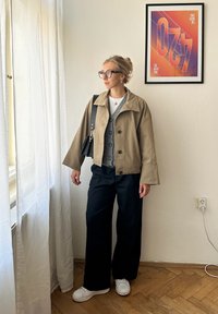 Lys brun cropped jakke med bred krage over en grå cardigan og hvit skjorte. Svart vide bukser og hvite sneakers fullfører looken.