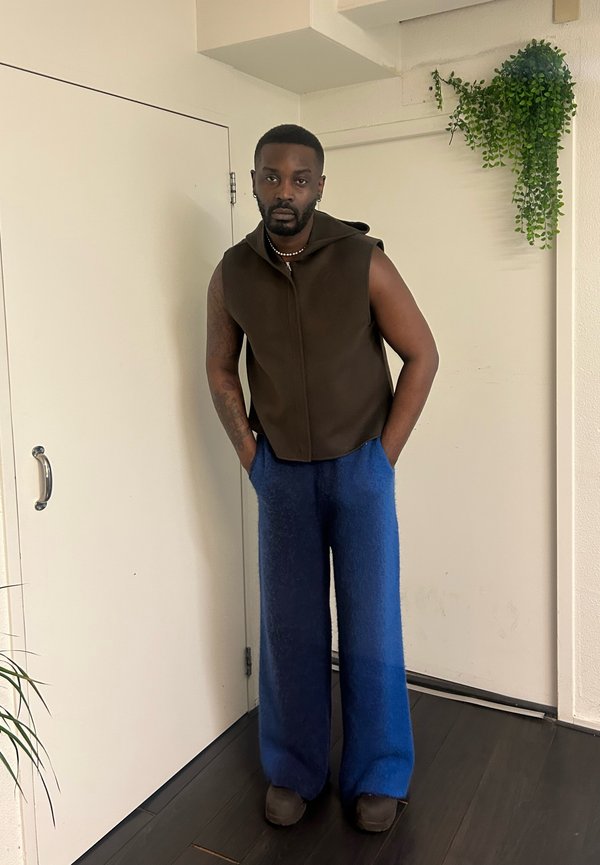 Chaleco marrón sin mangas con cremallera y capucha, combinado con pantalones anchos de textura azul, y calzado marrón. Entorno interior con vegetación.