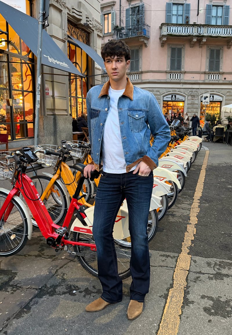 Mladý muž v rifľovej bunde a topánkach stojaci pri rade požičových bicyklov na mestskej ulici s obchodmi a ľuďmi v pozadí.