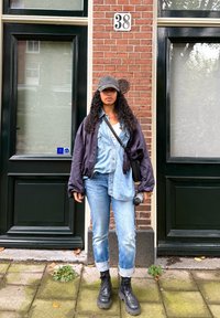 Individu in een zwarte bomberjack over een denim shirt, lichte spijkerbroek en zwarte chunky laarzen, met een camera in de hand, voor een steenmuur.