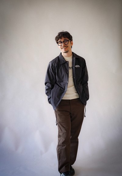 Chaqueta cortavientos negra con cierre a presión, llevada sobre una camiseta de cuello redondo de color claro, combinada con pantalones marrones y zapatillas negras.