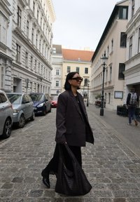 Tamsus, per didelį blizguną su pečių pagalvėlėmis, juoda suknelė ir didelė pintinė rankinė. Rudos saulės akinius ir kulkšnines batus užbaigia įvaizdį akmeninėje gatvėje.