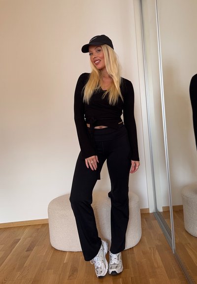 Top negro de manga larga con detalle de lazo, combinado con pantalones negros ajustados y zapatillas gris. En el fondo se ve un taburete de color claro y un espejo.