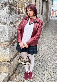 Bordo usnjena jakna, bela pletena srajca, krilo s zelenim karirastim vzorcem, bele nogavice, burgundski športni copati in potiskana torbica. Ozadje iz tlakovcev.