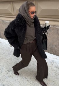 Kobieta ubrana w czarny futrzany płaszcz, brązowe spodnie i brązowy sweter z kapturem, trzyma filiżankę kawy i czarną torebkę, idąc po zaśnieżonym chodniku.