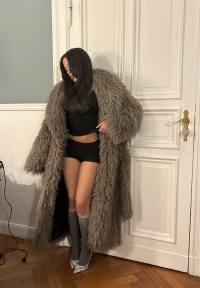 Mujer con un abrigo largo y desgreñado marrón sobre pantalones cortos y top negros, calcetas grises hasta la rodilla y zapatos de tacón a cuadros, de pie en el interior.