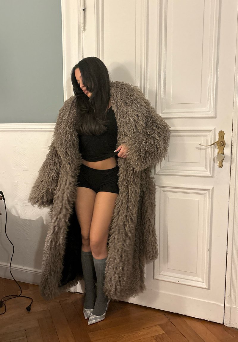 Frau, die einen langen, zotteligen braunen Mantel über schwarzer Shorts und Oberteil trägt, graue knielange Socken und karierte High-Heel-Schuhe, steht drinnen.