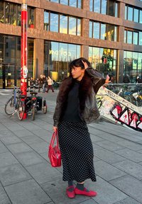 Falda negra con lunares, suéter negro, chaqueta de cuero con cuello de piel, bolso rojo y zapatos rojos. Entorno urbano con bicicletas y un edificio.