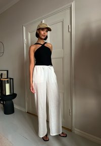 Črna rebrasta majica brez rokavov s križnimi naramnicami, bele lanene široke hlače in bež kapa. Črne japonke. Minimalističen notranji prostor.