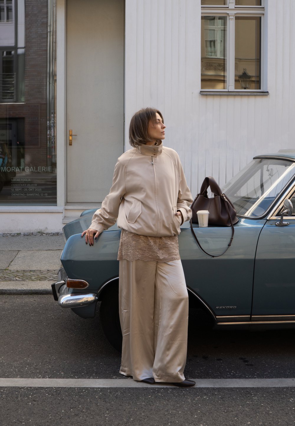 Giubbotto bomber beige, top in pizzo e pantaloni larghi in satin abbinati a una borsa marrone. In piedi accanto a un'auto vintage blu su una strada cittadina.