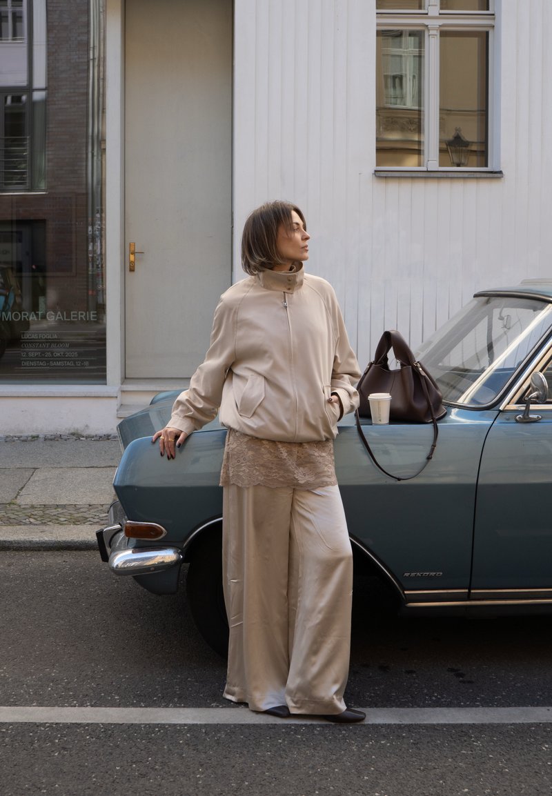 Giubbotto bomber beige, top in pizzo e pantaloni larghi in satin abbinati a una borsa marrone. In piedi accanto a un'auto vintage blu su una strada cittadina.