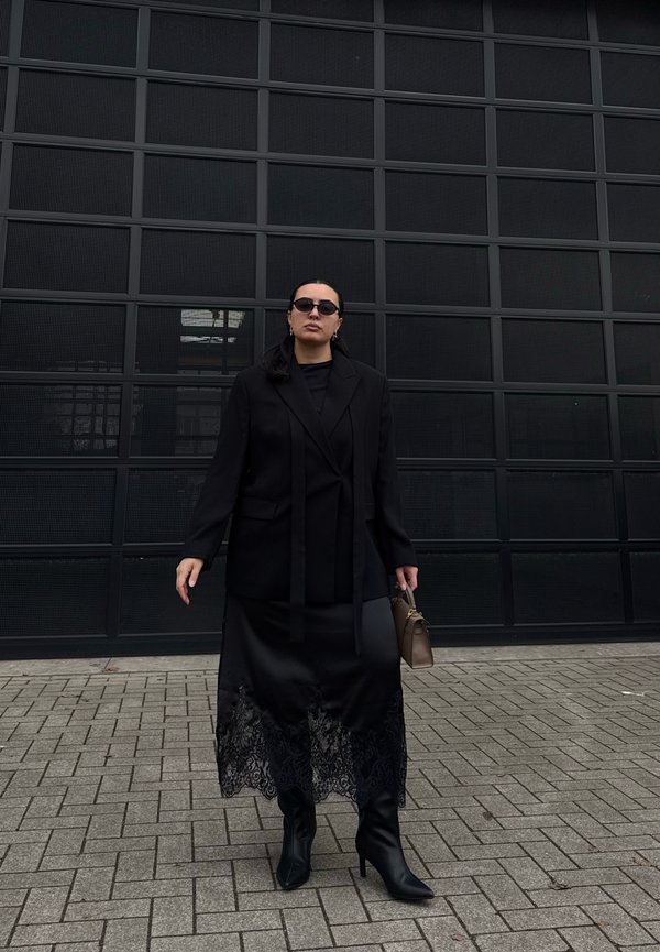 Žena v čiernom saka, čiernej sukni s čipkovým lemom, s koženými čižmami, slnečnými okuliarmi a zlatými kruhovými náušnicami, nesie malú hnedú kabelku a stojí na chodníku.