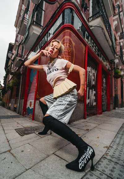 Bolso plisado beige, camiseta blanca de manga corta con estampado, falda metálica plateada, botas altas negras con texto blanco, entorno urbano.
