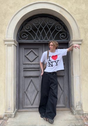 Camiseta blanca con estampado negro "I ♥ NY", pantalones de pierna ancha negros y un bolso de hombro beige. El fondo presenta una puerta de madera texturizada.