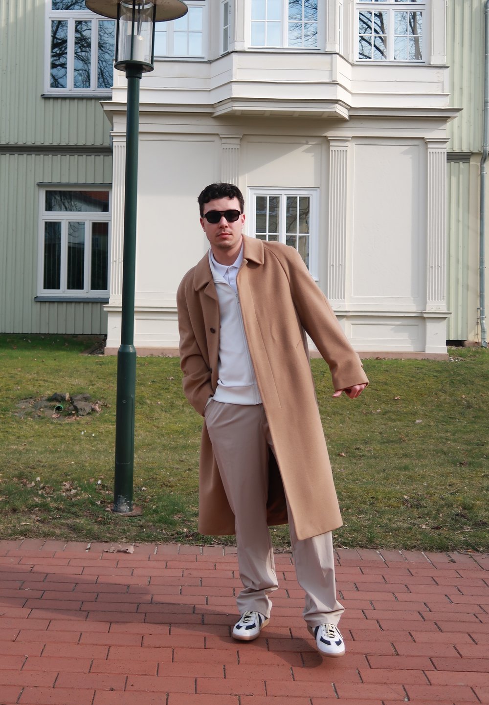 Beige overjas met een relaxte pasvorm, gedragen over een witte poloshirt en beige broek. Zwarte zonnebril en sportieve schoenen maken de look compleet.