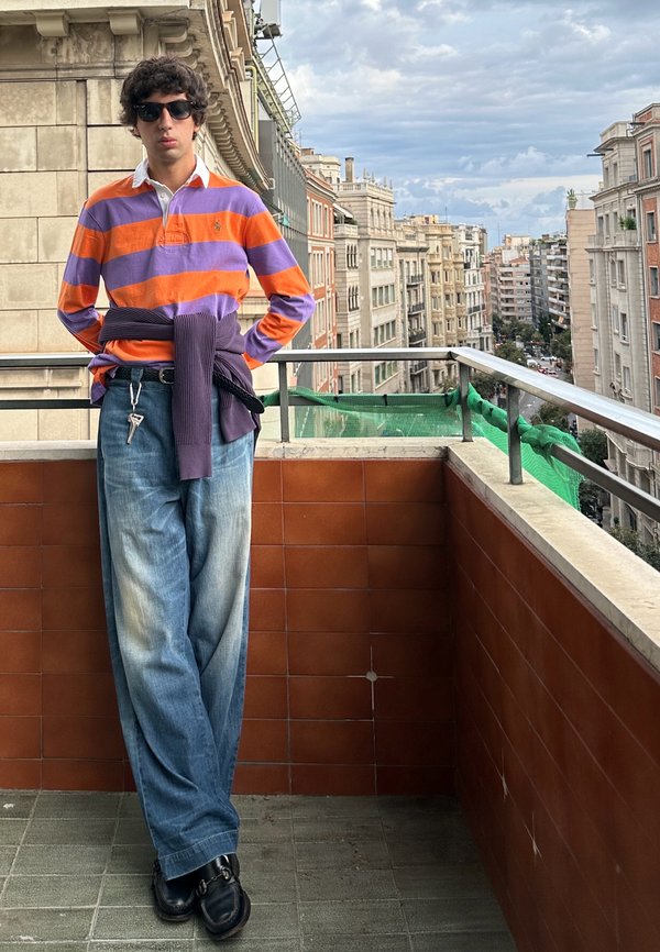 Persona joven con gafas de sol, una camisa de rayas moradas y naranjas y vaqueros holgados, de pie en un balcón con vista a una calle de la ciudad con edificios altos.