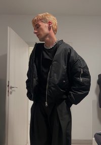 Jachetă bomber neagră cu mâneci supradimensionate, asortată cu o cămașă neagră. Colier din lanț argintiu notabil �și accesorii pentru urechi. Fundal neutru.