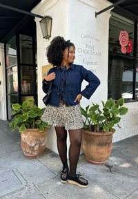 Dunkelblaue Jeansjacke mit vorderen Bindebändern über einem Leopardenmuster ausgestellten Rock. Schwarze Plateauschuhe und transparente schwarze Strumpfhosen. Topfpflanzen im Hintergrund.