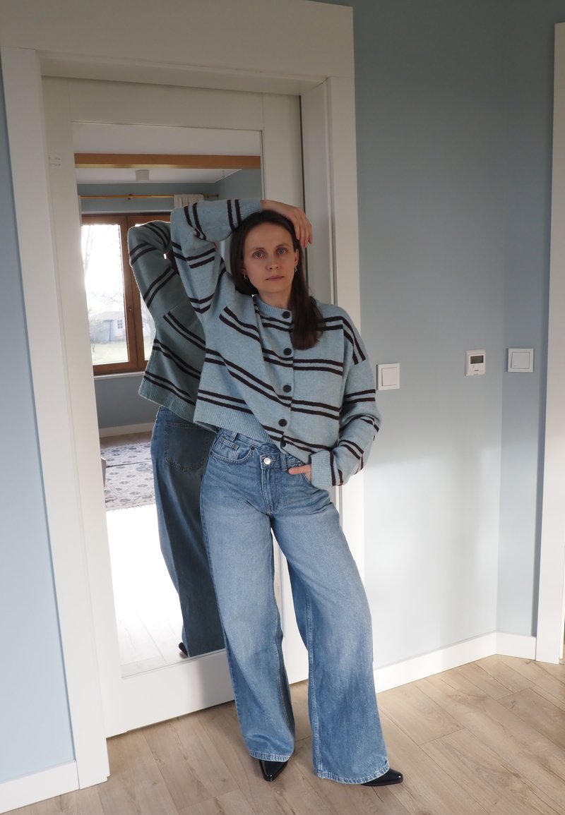 Pull court bleu clair avec des rayures foncées et des boutons, associé à un jean taille haute à jambes larges. Un miroir reflète la même tenue.