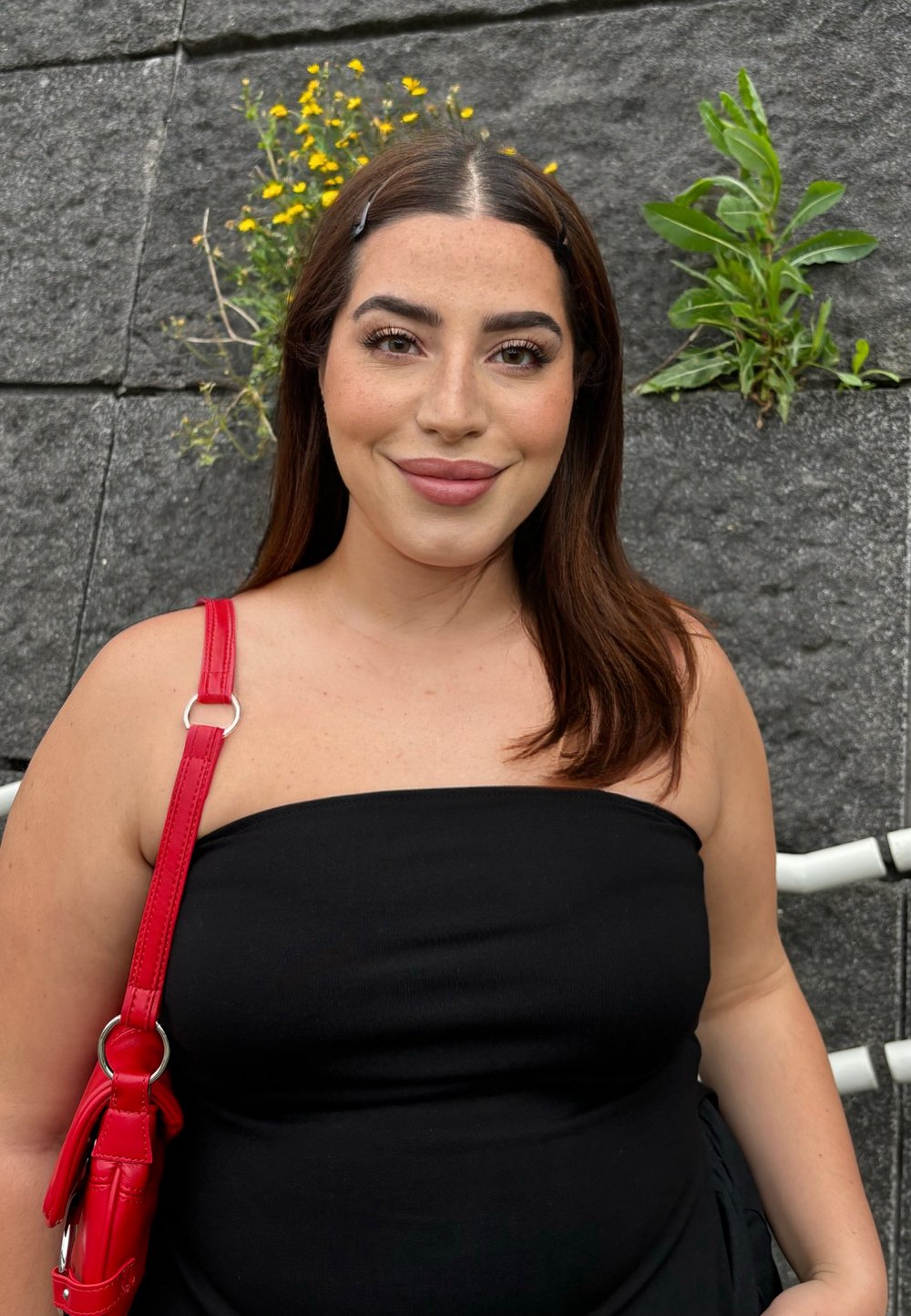 Svart, axelbandslös topp i kombination med en röd handväska med silverringdetalj; medellångt brunt hår med dekorativa klipp; bakgrund av stenmur.