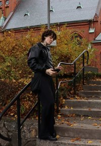 Ung person med krøllet hår og sort læderjakke, der lytter til musik, mens de bruger smartphone på udendørs trapper med efterårsblade og murstensbygning.